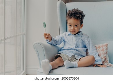 Portrait of cute adorable little boy with big eyes holding round blue sweet and tasty lollipop and looking at camera, casually dressed sitting alone on comfy armchair in minimalist room - Shutterstock ID 2003873204