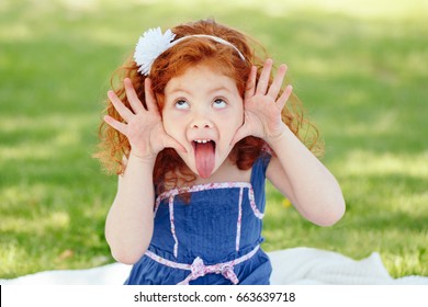 Portrait of cute adorable little red-haired Caucasian girl child in blue dress making funny silly faces showing tongue, in park outside, playing  crying screaming, having fun, lifestyle childhood