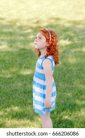 Portrait Of Cute Adorable Little Red-haired Caucasian Girl Child In Blue Dress Standing In Field Meadow Park Outside, Looking Up In The Air, Having Fun, Happy Lifestyle Childhood Concept