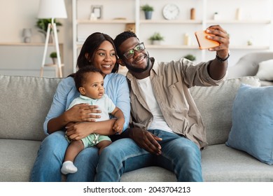 Portrait Of Cute African American Dad And Mom Taking Selfie With Their Kid At Home, Cheerful Black Family Having Good Time Together, Taking Photos On Smartphone Camera, Sitting On Couch In Living Room