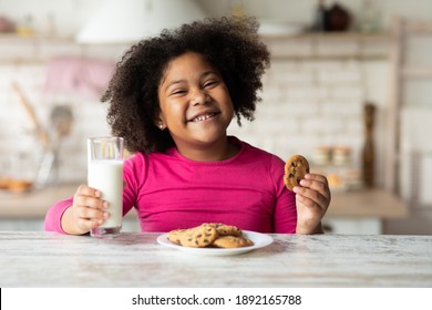 Portrait Of Cute African American Girl Enjoying Cookies And Milk In Kitchen, Pretty Black Child Eating Snack At Home, Preschool Kid Having A Bite With Fresh Baked Biscuits And Calcium Drink, Closeup