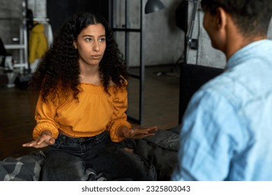 Portrait of cute african american girl of 20s with long curly hair sitting on bed in yellow top, gesticulating while telling something to her boyfriend turned back to camera, planning weekend - Powered by Shutterstock