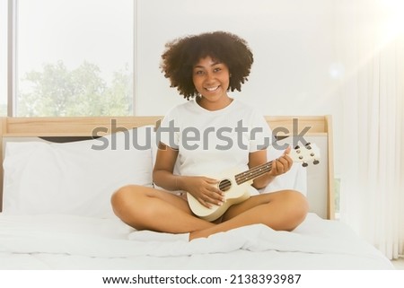 Portrait cute african american woman lounging and practicing her ukulele in bed with pleasure : Living a lifestyle and having fun at home concept.
