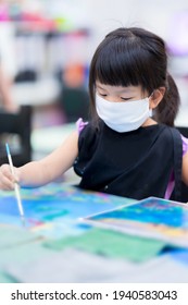 Portrait Of Cute Asian Girl Wearing A White Cloth Face Mask. Students Are Studying Art In Class. Happy Children Paint The Water On The Canvas. Child Wears A Black Apron. Kids Is 4 Years Old.