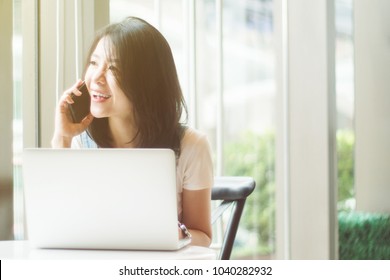 Portrait Of Cute Asian Teen Woman Talking Smartphone And A Laptops Or Notebook Computer In Coffee Cafe. Morning Or Sunset Moment. Filtered Image And Light Effect Added. Happy Smile Chill Out Concept