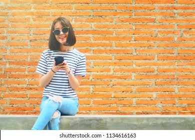 Portrait Of Cute Asian Teen Woman Using Smartphone At Outdoor. Texting With Someone. Reading Pleasant Text Message On Mobile Phone While Happy Smile Chill Out Place. Technology Communication Concept