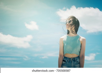 Portrait Of A Cute Asian Teenager Young Woman Stand Against Blue Sky And Cloud Background. Wearing Sunglasses. Wind Blows Her Hair. Filtered Image To Vintage Tone. Light Effect Added. Hipster Concept