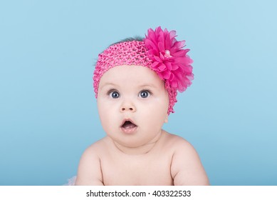 Portrait Of Cute Baby With Pink Bow Flower On Her Head. Surprised Baby Child Looking Into The Camera. Lovely Kid With Big Blue Eyes. Happy Family Concept