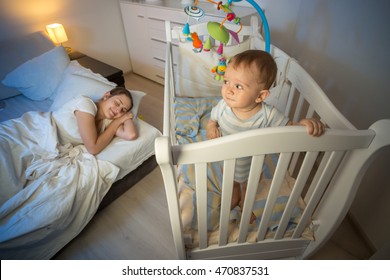 Portrait Of Cute Baby Standing In Crib And Looking At Tired Mother That Fell Asleep