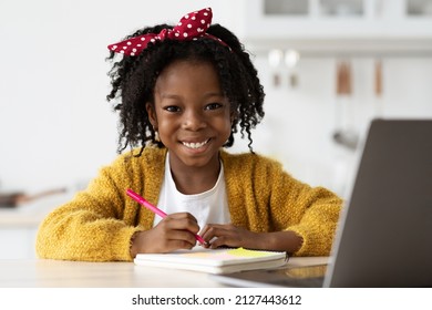 Portrait Of Cute Black Girl Writing With Pen In Notepad And Smiling At Camera While Sitting At Desk At Home, Happy Cheerful African American Female Child Doing School Homework, Closeup Shot - Powered by Shutterstock