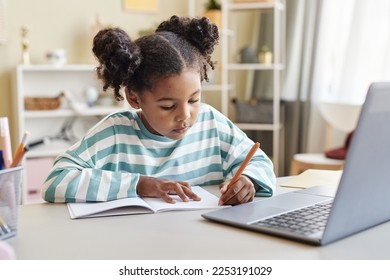 Portrait of cute black girl writing in notebook while doing homework at desk in minimal home interior - Powered by Shutterstock