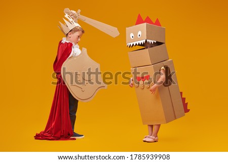Portrait of a cute boy in costume of knight with cardboard armour is fighting the cardboard dragon. Childhood dreams. Full length portrait on a yellow background.