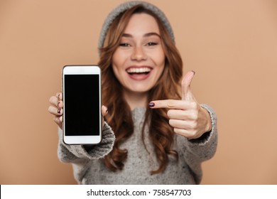 Portrait of cute brunette girl in warm hat and sweater pointing with finger on blank mobile screen, selective focus - Powered by Shutterstock