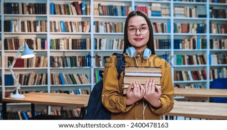 Portrait of cute brunette student with white headsets, black backpack and lots of book in hands smiling and looking in camera in library.