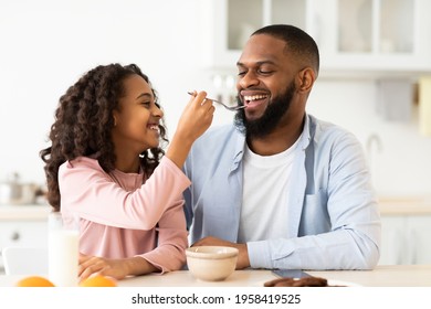 Portrait Of Cute Caring African American Little Girl Feeding Her Happy Father With Cereal While They Having Breakfast At Home In The Kitchen. Bonding, Love, Paternity And Family Concept