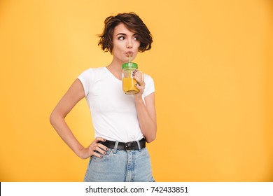 Portrait of a cute casual girl drinking orange juice from a glass and looking at camera isolated over yellow background