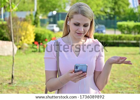Portrait of a cute caucasian girl outdoors talking on a smartphone. Pretty blonde model posing while standing in sunny weather in a city park.