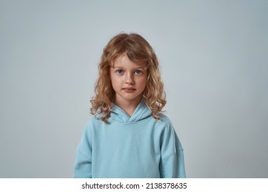 Portrait Of Cute Caucasian Little Girl Looking At Camera. Cropped Image Of Serious Blonde Curly Female Child Wearing Hoodie. Childhood Concept. Isolated On Grey Background. Studio Shoot. Copy Space