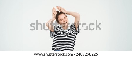 Portrait of cute caucasian woman, makes animal ears, cute pose, smiling at camera, standing over white background.