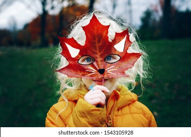 Portrait Of Cute Child Kid With Blue Eyes Holding Autumn Fall Red Maple Leaf Painted Mask In Front Face. Girl Playing Pretending To Be Fox Cat. Fairy Dreamy Authentic Childhood. Halloween Holiday.