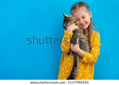 Portrait of a cute child who with tenderness and love hugs a gray cat in his arms and smiles with happiness on a blue background. The concept of pets.