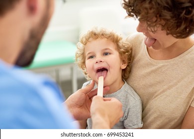 Portrait Of Cute Curly Child Sitting On Mothers Lap During Medical Checkup, With Doctor Inspecting His Open Mouth