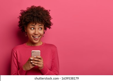 Portrait of cute curly haired girl scrolls social networks or does shopping online, looks aside positively, uses modern smartphone, wears pink sweater, poses indoor in studio, reads message.