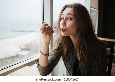 Portrait of cute eating woman with spoon sitting near window in cafe