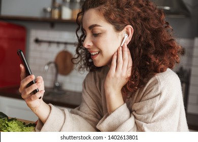 Portrait of cute european woman with earpods listening to music on cellphone while cooking in kitchen interior at home