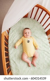 Portrait of cute  funny adorable white Caucasian little baby newborn in yellow onesie lying in crib alone near window, lifestyle candid real life, view from top above