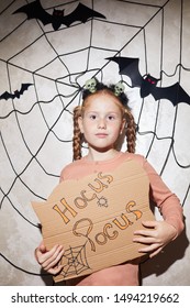 Portrait cute girl holding Hocus Pocus placard   posing at camera and cobweb   bats in the background