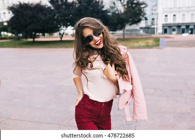 Portrait of cute girl with long curly hair and phone in hands smiling to camera in city on bulding background.