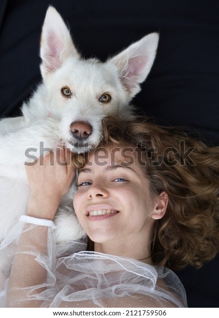 portrait of a cute\
girl - a teenager of 17-18 years old next to her white dog on a\
dark background. the similarity and friendship between the female\
owner and pet. world dog\
day