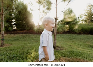 Portrait Of Cute Handsome Blond Little Kid Boy In Colorful Striped Button Down Shirt Walking Outside In The Grass Watching The Sunrise