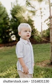 Portrait Of Cute Handsome Blond Little Kid Boy In Colorful Striped Button Down Shirt Walking Outside In The Grass Watching The Sunrise