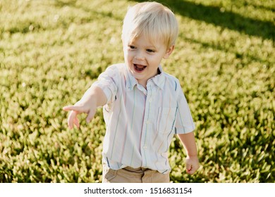 Portrait Of Cute Handsome Blond Little Kid Boy In Colorful Striped Button Down Shirt Walking Outside In The Grass Watching The Sunrise