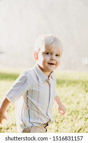 Portrait Of Cute Handsome Blond Little Kid Boy In Colorful Striped Button Down Shirt Walking Outside In The Grass Watching The Sunrise