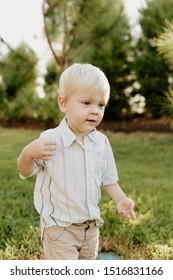 Portrait Of Cute Handsome Blond Little Kid Boy In Colorful Striped Button Down Shirt Walking Outside In The Grass Watching The Sunrise