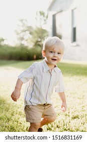 Portrait Of Cute Handsome Blond Little Kid Boy In Colorful Striped Button Down Shirt Walking Outside In The Grass Watching The Sunrise