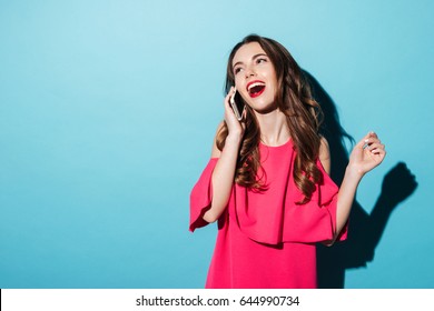 Portrait Of A Cute Happy Girl In Dress Talking On Mobile Phone And Laughing Isolated Over Blue Background