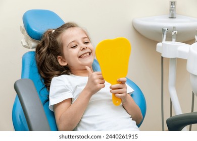 Portrait of a cute happy little girl sitting on a chair in a dental office, showing thumbs up after a dental examination while holding mirror in her hand. - Powered by Shutterstock