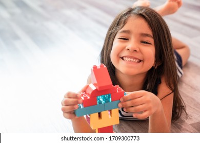 Portrait of a cute happy little preschool Hispanic girl playing alone with colorful construction blocks in her bedroom floor at home. Child Developing Motor Skills With Games Copy Space