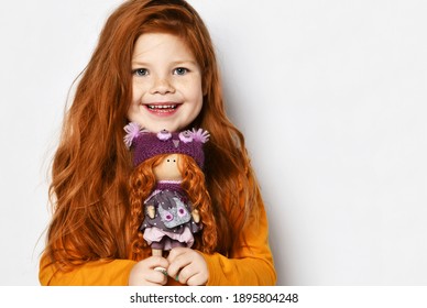 Portrait of cute happy smiling red-haired kid girl in orange sweatshirt holding showing small redhair doll her portrait in hands at face over white background with copy space