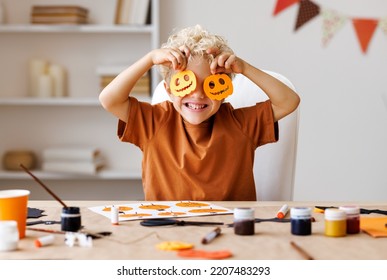 Portrait of cute kid  boy  making Halloween home decorations   while sitting at wooden table, child covers eyes with carved pumpkins and laughs merrily