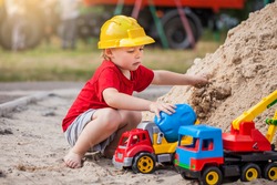 Portrait Of Cute Kid In Construction Helmet Sitting On Sand And Playing With Tractor And Construction Toys. Child Walks Outdoor. Life Style