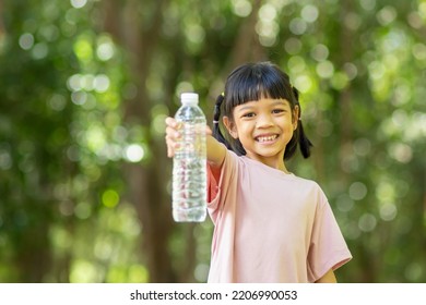 Portrait, A Cute  Kid Girl, Age 6 Years And Over, Wearing A Pastel Pink Shirt. Standing Water Bottle With Her Right Hand Outstretched In Front With A Smiling And Happy Face. In A Shady Green Park. 