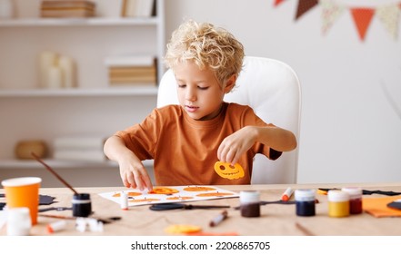 Portrait Of Cute   Kids Boy  Making Halloween Home Decorations Together While Sitting At Wooden Table, Child Painting Pumpkins And Making Paper Cuttings