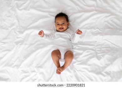 Portrait Of Cute Laughing Black Infant Baby Lying On Bed, Adorable Little African American Child In White Bodysuit Relaxing In Bedroom And Looking At Camera, Top View Shot With Copy Space - Powered by Shutterstock