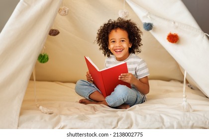 Portrait of cute little african american kid curly boy with book smiling at camera while   reading book  in play tent at home, happy child playing alone in children room - Powered by Shutterstock