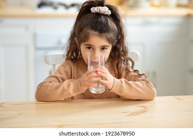 Portrait Of Cute Little Arab Girl Drinking Water From Glass, Pretty Female Child Sitting At Table In Kitchen And Enjoying Healthy Mineral Drink, Preschooler Kid Looking At Camera, Closeup, Free Space